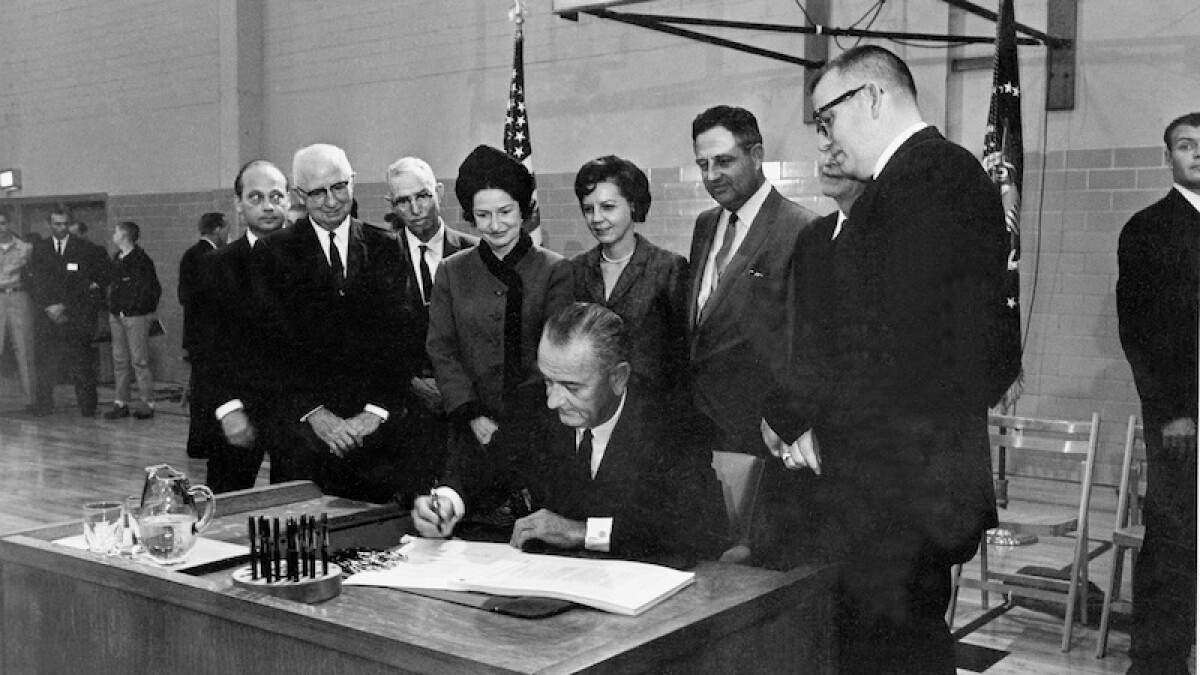 A group of men crowd a man sitting at his desk who is signing a document