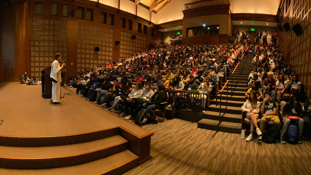 Presenter is standing centerstage while facing a full auditorium of students listening to their speech/presentation